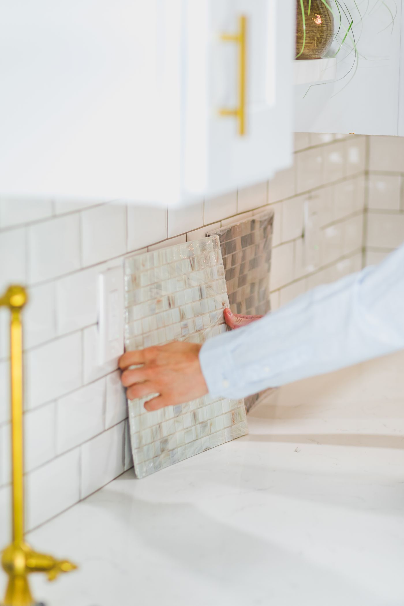 A hand holding a mosaic tile sample flat against an installed tile wall to compare the two.