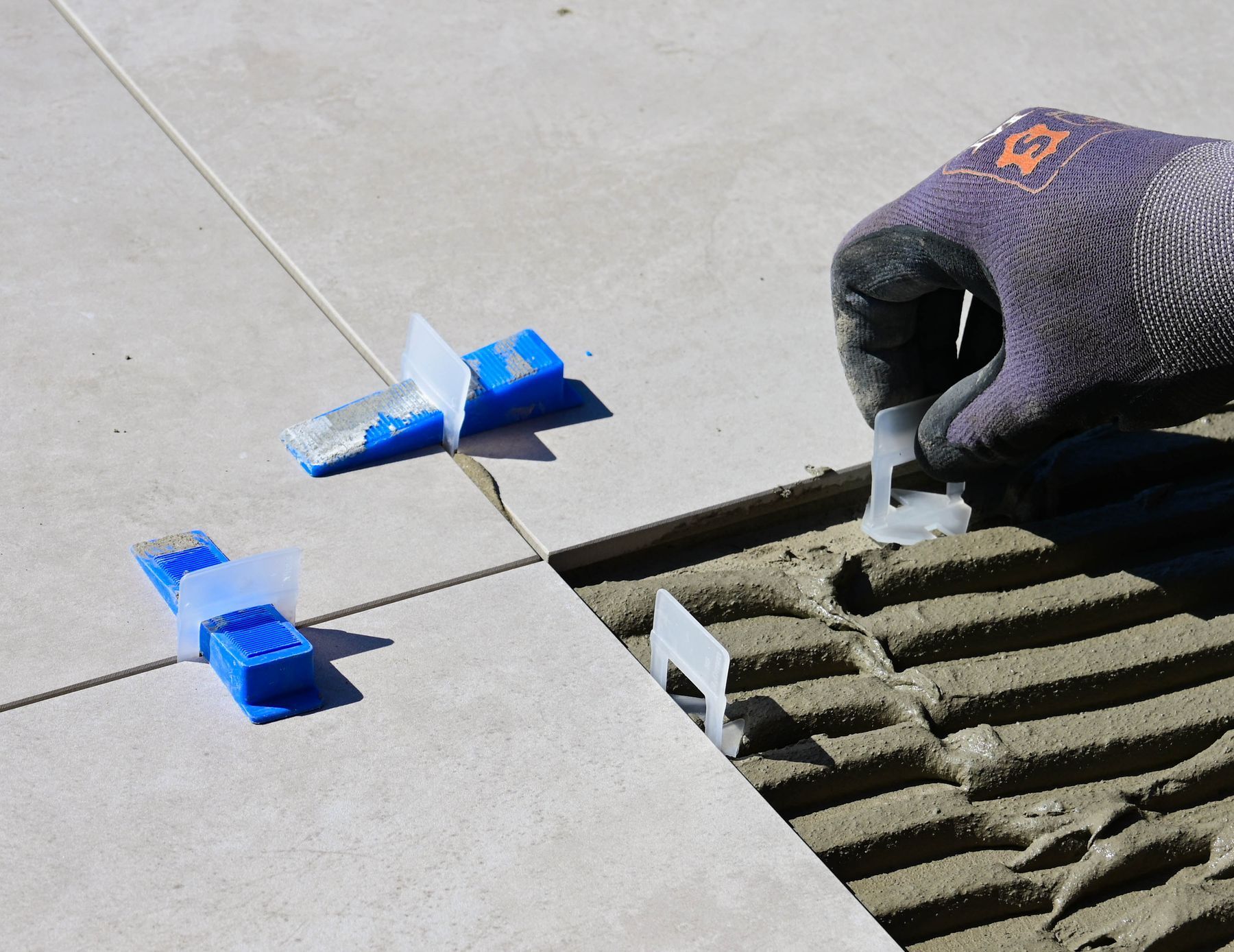 A gloved hand setting a tile into combed thinset, with levelling clips holding the last row flat.