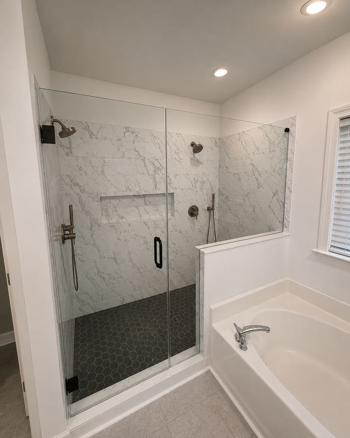 The finished shower: marble-look subway tile to the ceiling, a recessed niche, black hex mosaic floor and a frameless glass door.